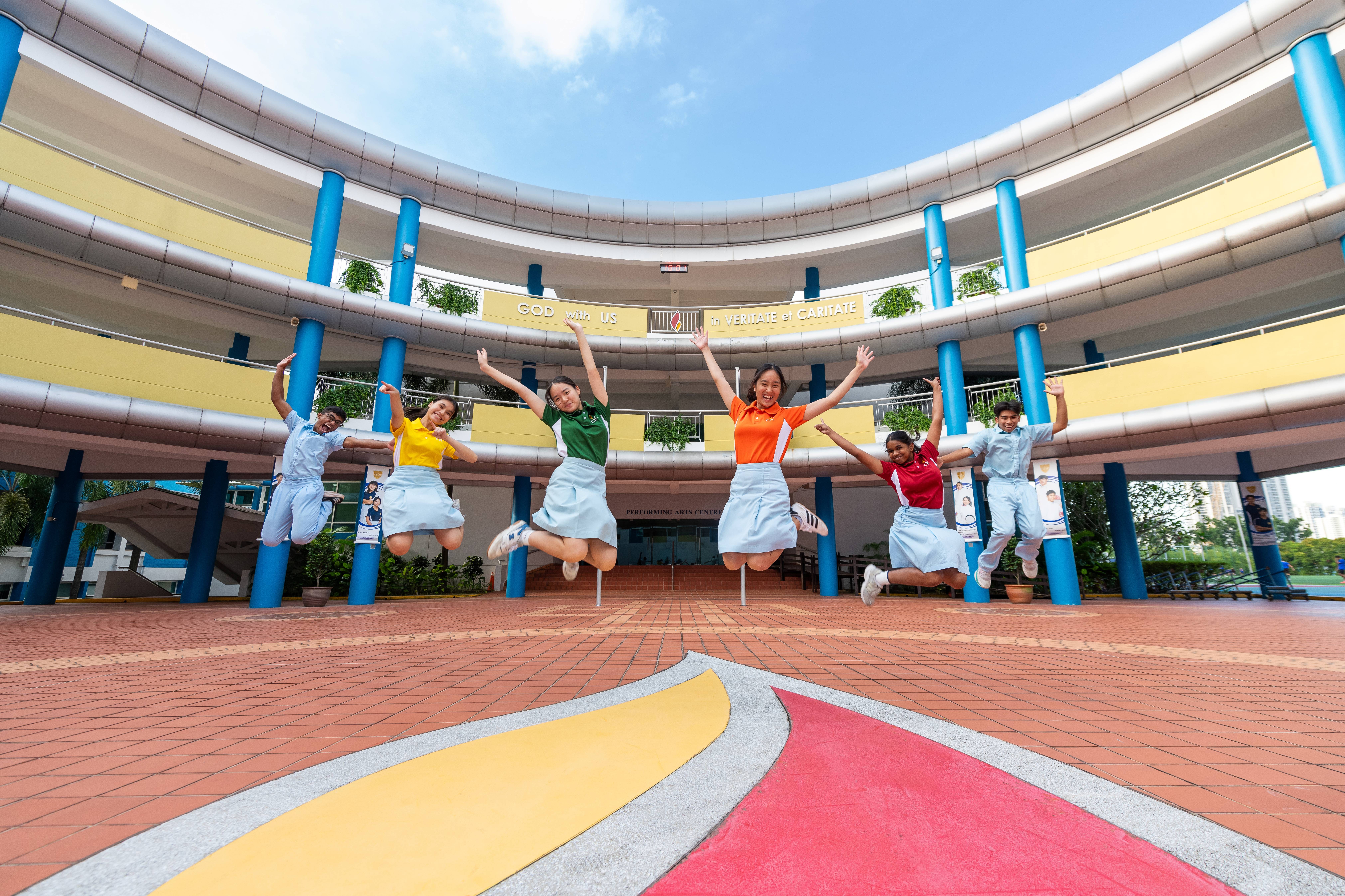 Four smiling CJC students in red, yellow, orange, and green uniforms walk and chat on a school field.